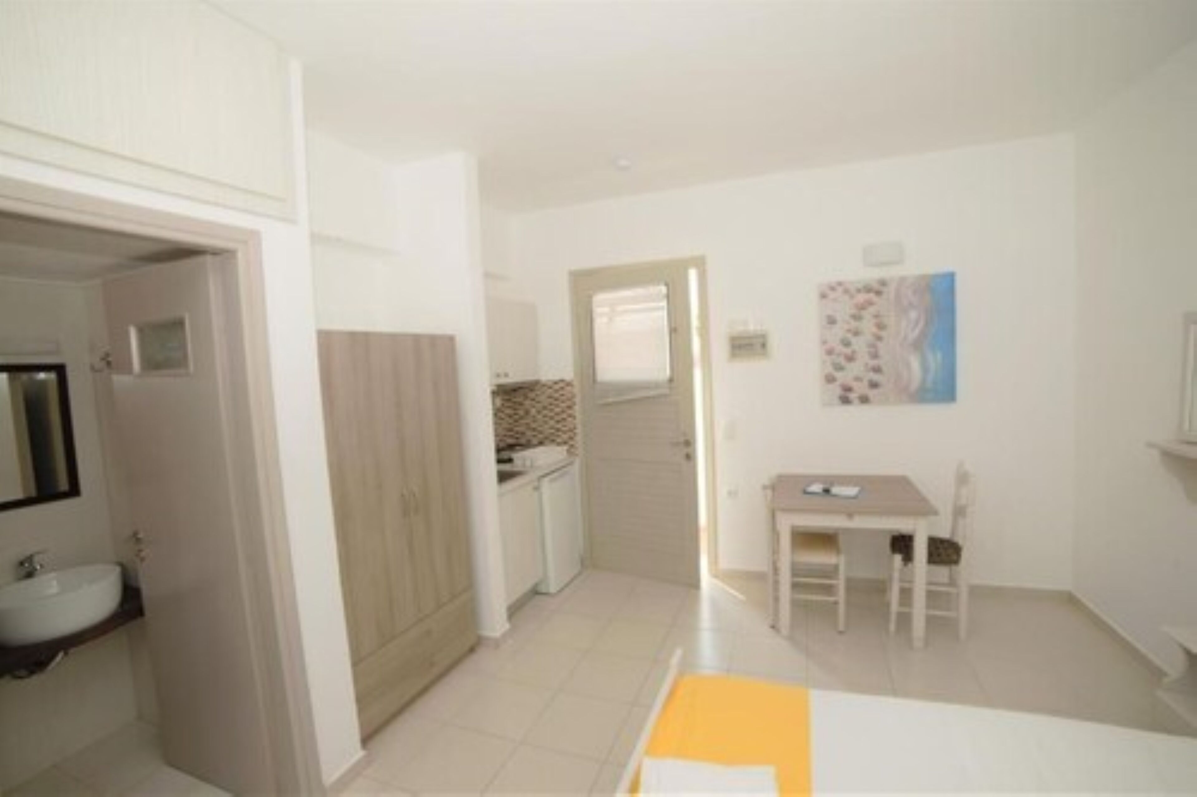 A small, bright room with a white sink, wooden cabinets, a small table, and a painting on the wall.