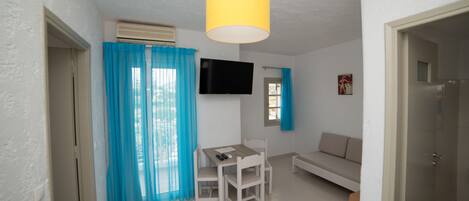A living room with a white sofa, a dining table with chairs, a TV mounted on the wall, and a yellow hanging light fixture.