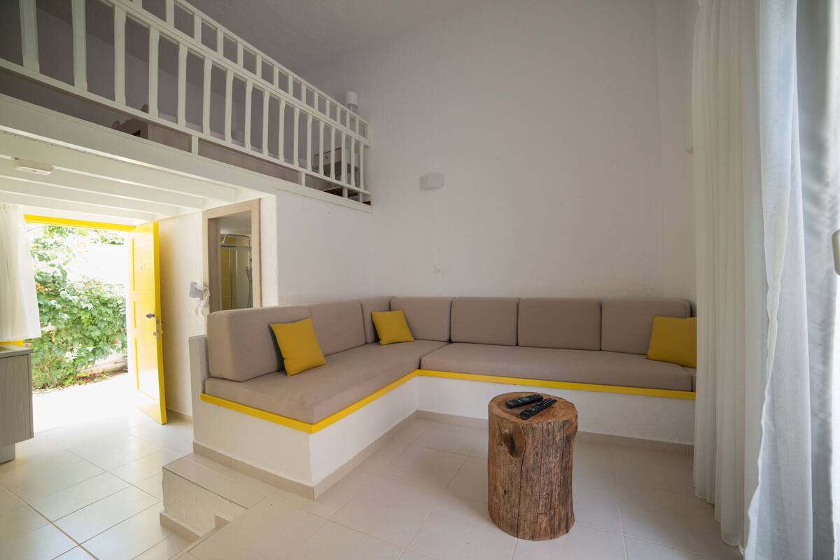 A modern living room with a beige sectional sofa, yellow accents, a wooden stool, and a staircase leading to an upper level.