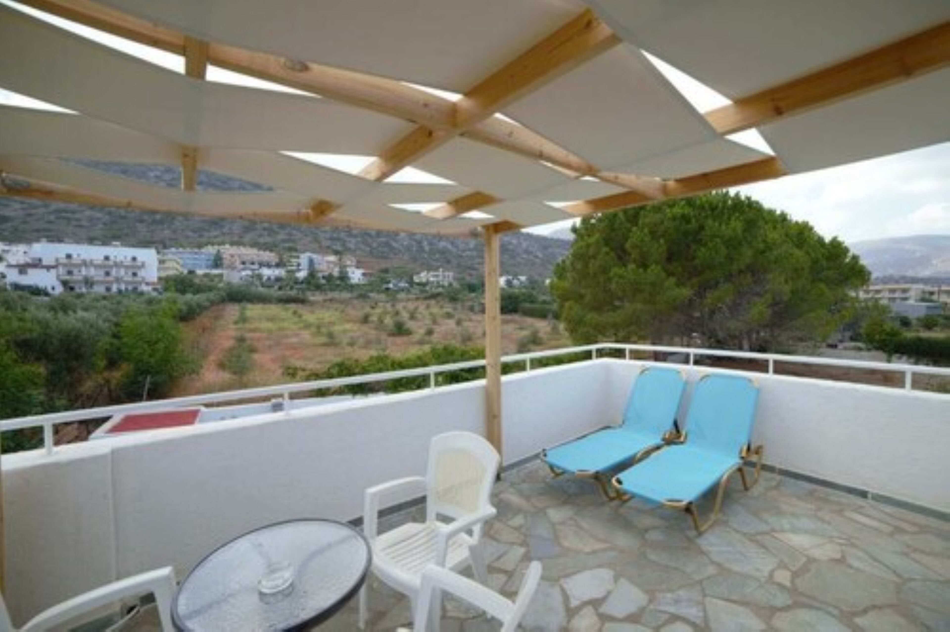 A balcony with a table and chairs, a wooden pergola, and a view of a hillside with buildings.