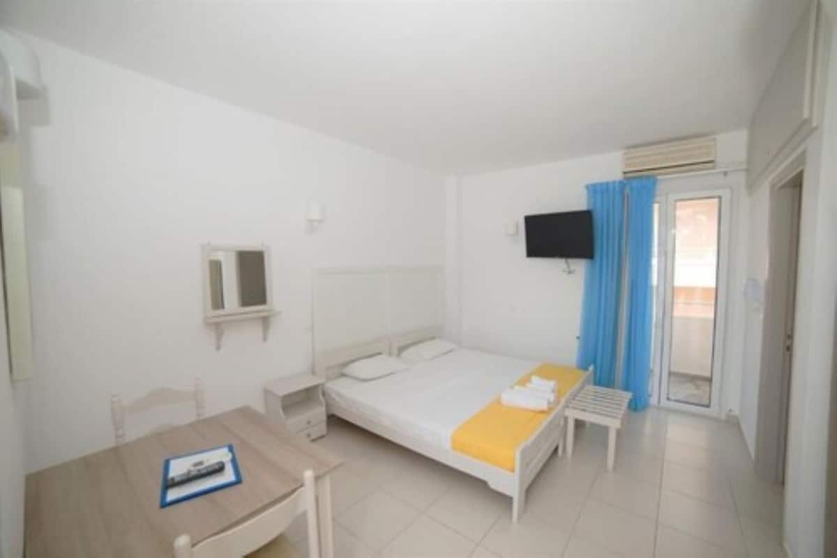 A bedroom with a bed, a desk, a chair, a TV mounted on the wall, and a window with blue curtains.