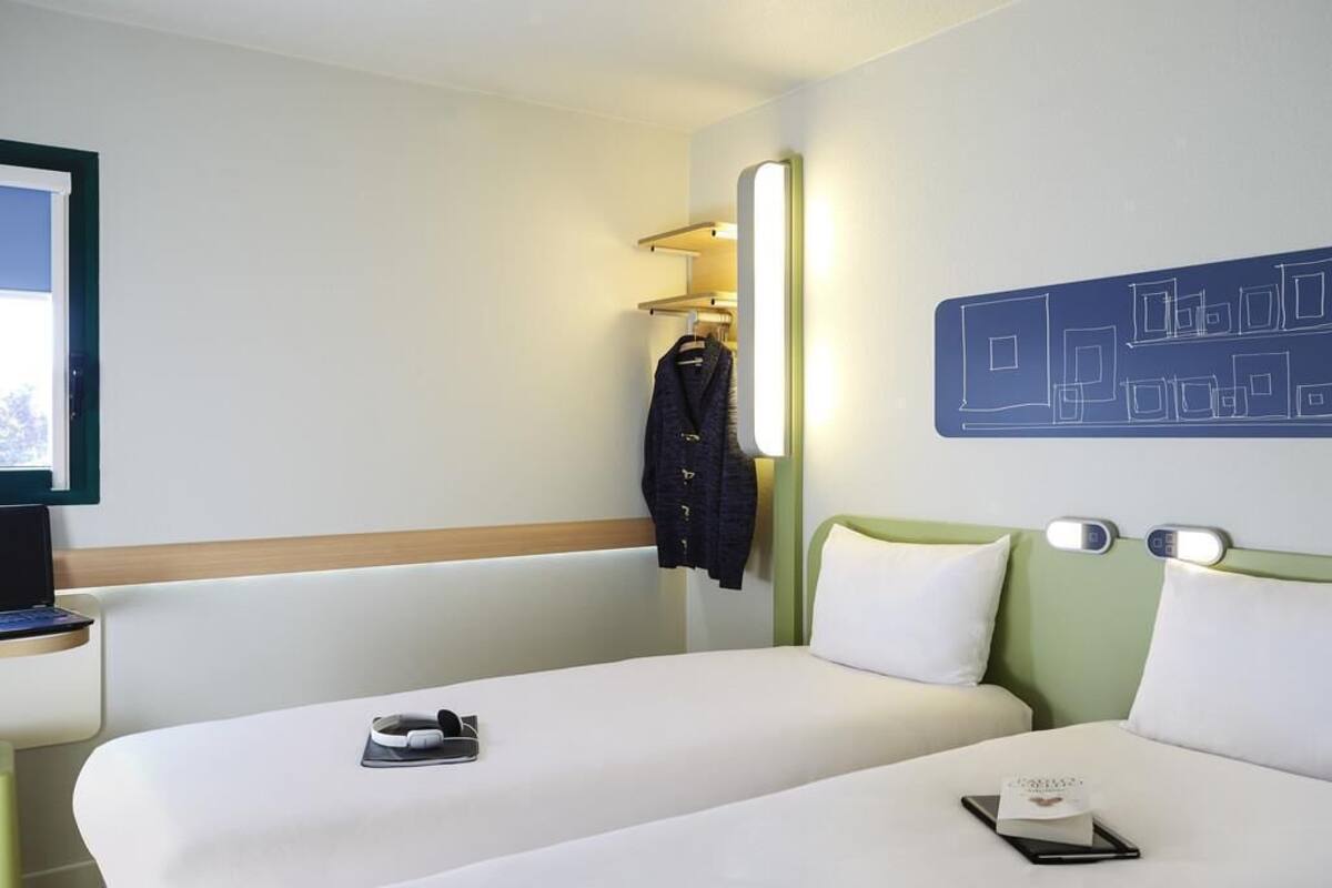 Ein Hotelzimmer mit zwei Betten, einem Schreibtisch mit einem Laptop, einem Kleiderständer und einer Wand mit geometrischem Muster.