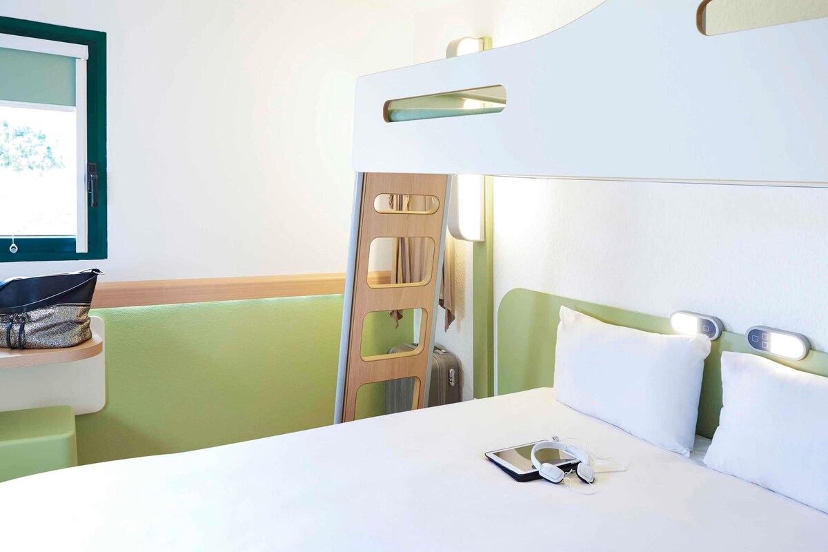 Una cama litera con una mesita de noche, un televisor en la pared y una maleta en el suelo.