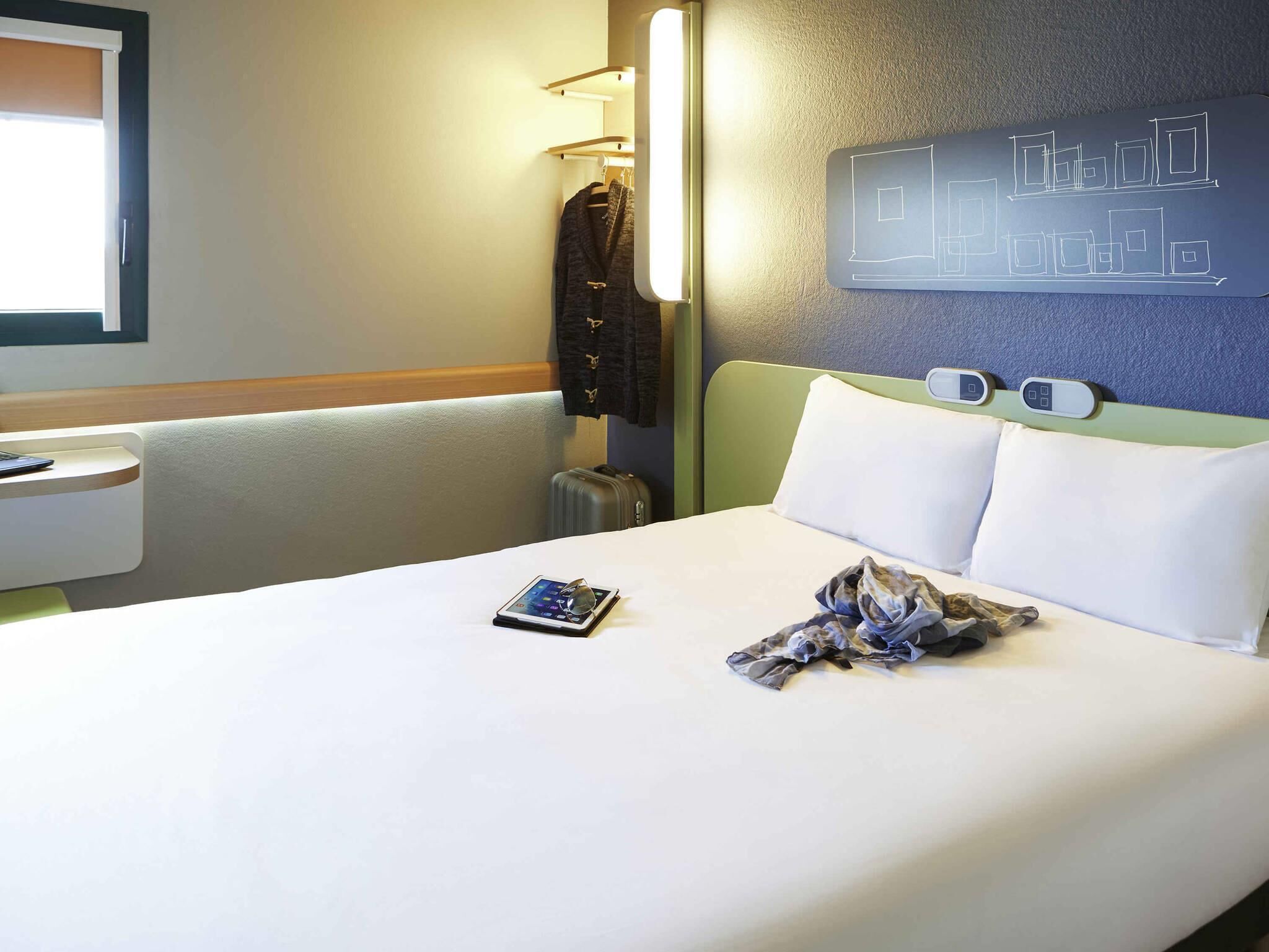 A hotel room with a bed, a bedside table, a lamp, a wall with a geometric design, and a window.