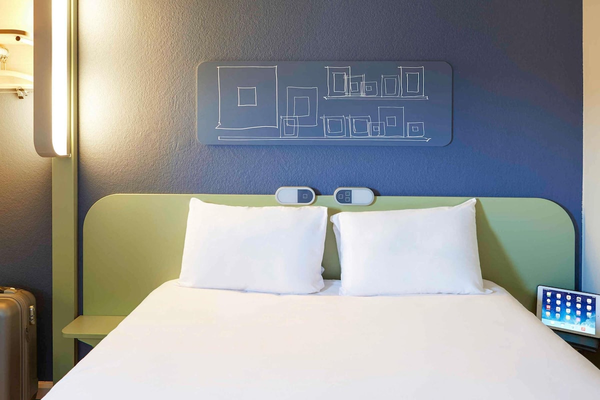 Une chambre d’hôtel avec un lit, un thermostat fixé au mur et une table de chevet avec une lampe.