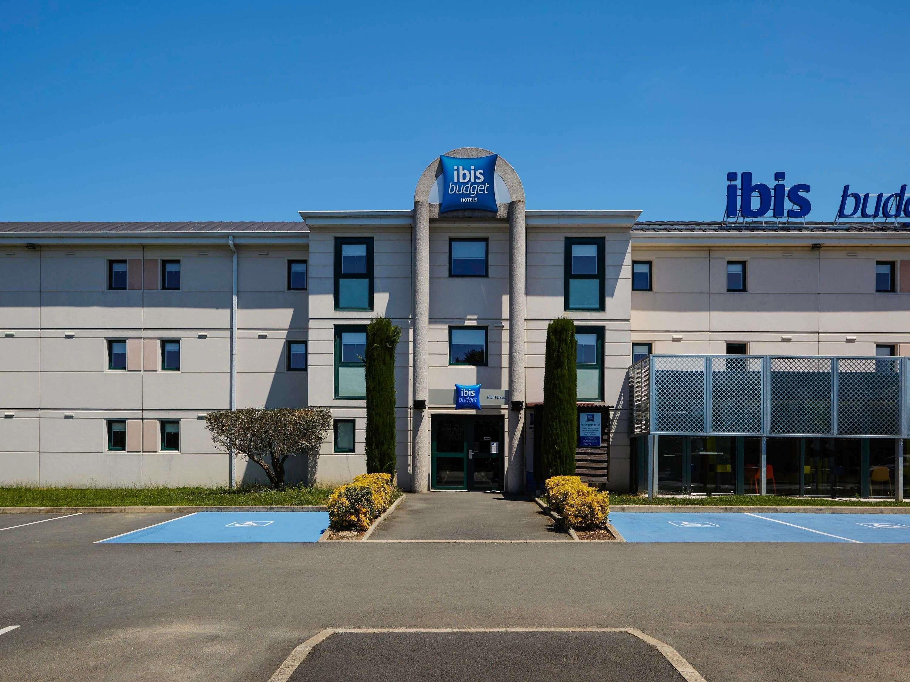 A building with 'ibis budget hotels' signage, a parking lot with blue handicapped parking spaces, and a clear sky.