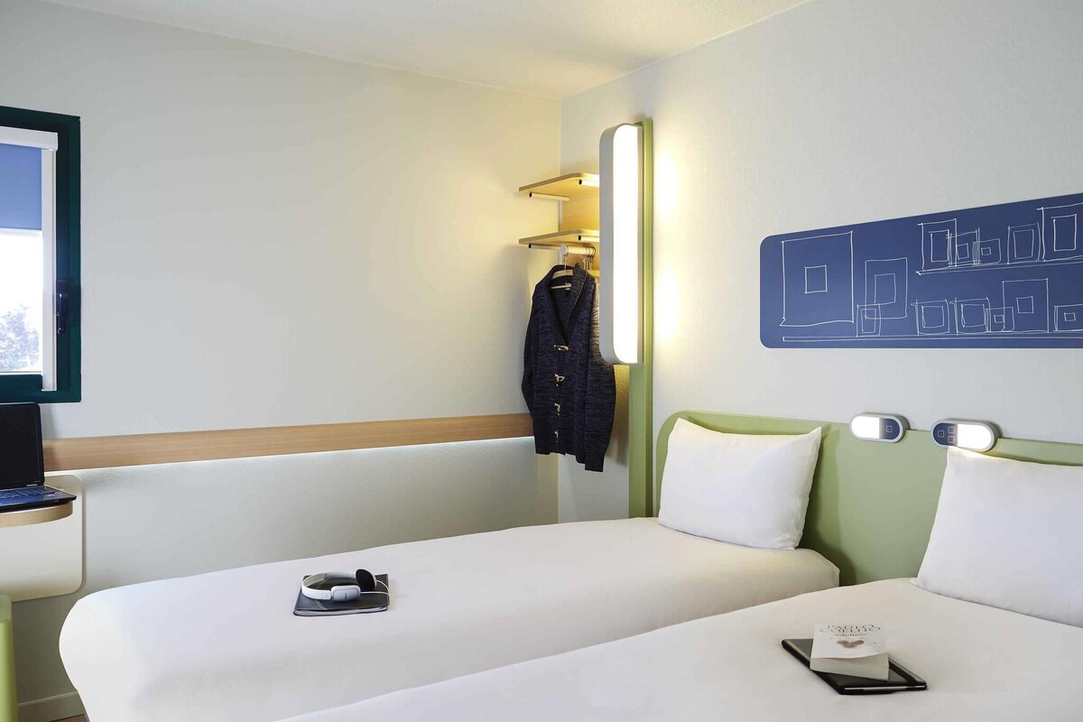 A hotel room with two beds, a desk with a laptop, a coat rack with a jacket, and a wall art piece.