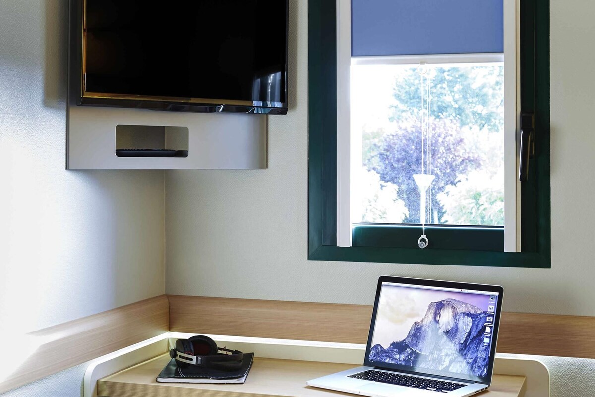 Une pièce avec un ordinateur portable sur un bureau, un téléviseur fixé au mur et une fenêtre donnant sur des arbres.