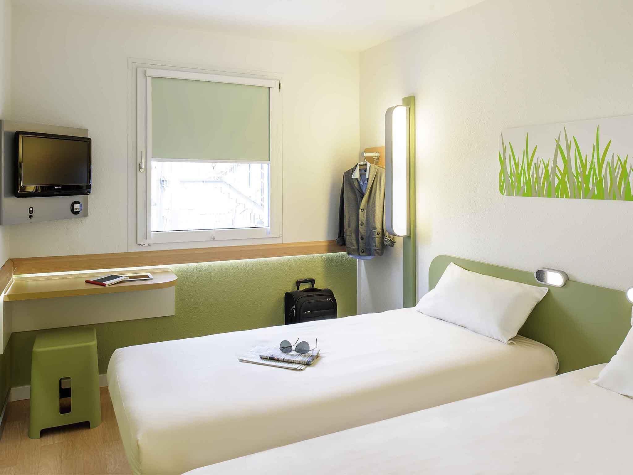 A hotel room with a bed, a desk with a TV, a window, and a wall with a green grass pattern.
