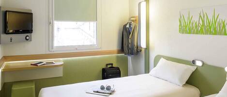 A hotel room with a bed, a desk with a TV, a window, and a wall with a green grass pattern.
