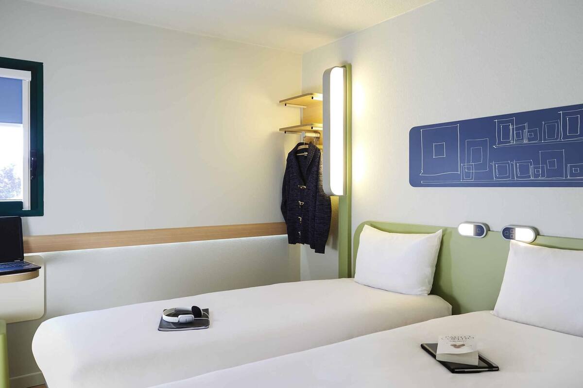 Une chambre d’hôtel avec deux lits, un bureau avec un ordinateur portable, un porte-manteau avec une veste, et une œuvre d’art murale.