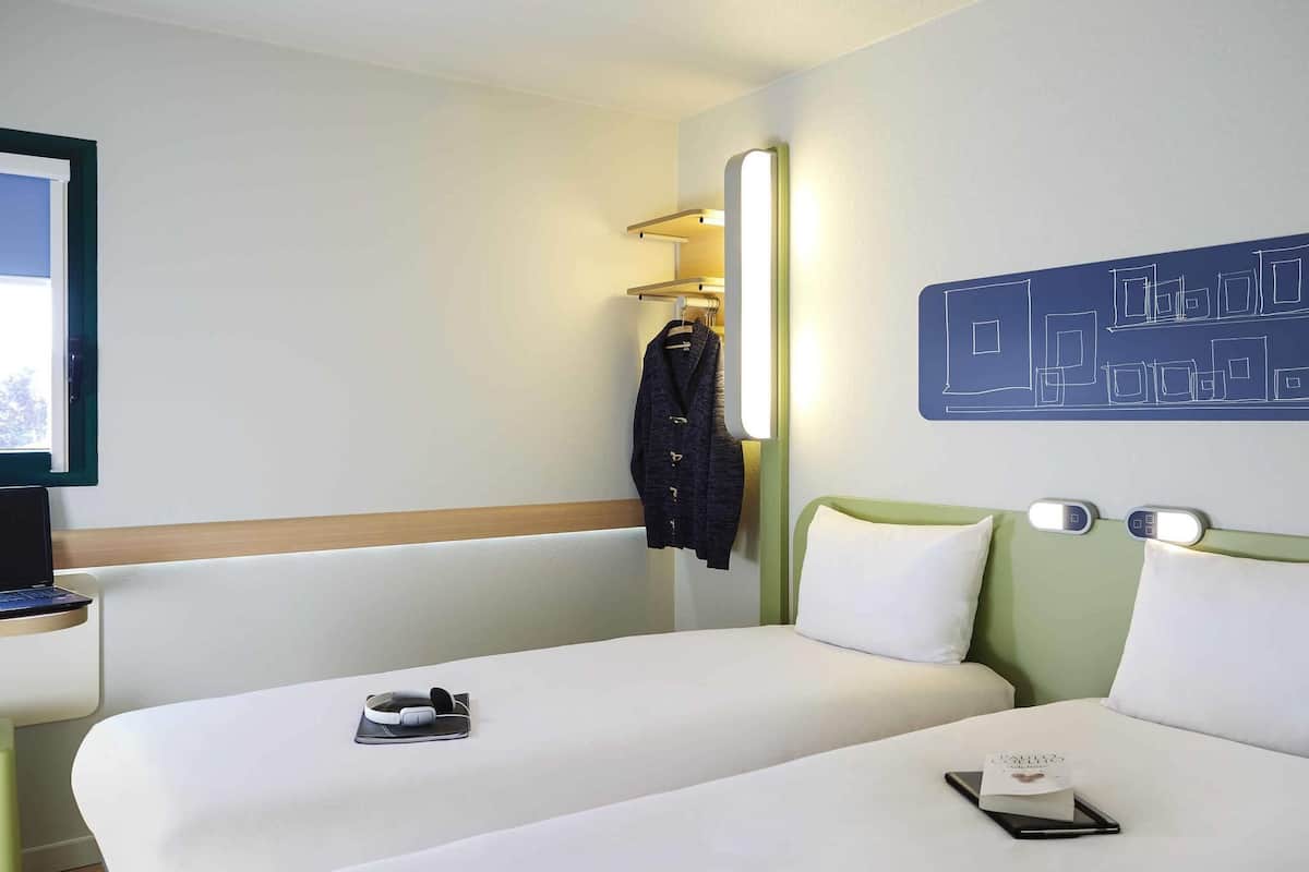 Hotellihuone, jossa on kaksi sänkyä, työpöytä kannettavalla tietokoneella, naulakko takille ja seinälle ripustettu taideteos.