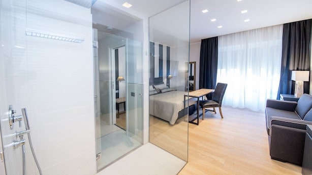 Habitación de hotel moderna con un separador de vidrio que divide el baño y el dormitorio. La habitación cuenta con una cama, un escritorio y un sofá.