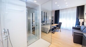 Een moderne hotelkamer met een glazen scheiding tussen de badkamer en slaapkamer. De kamer is voorzien van een bed, een bureau en een bank.