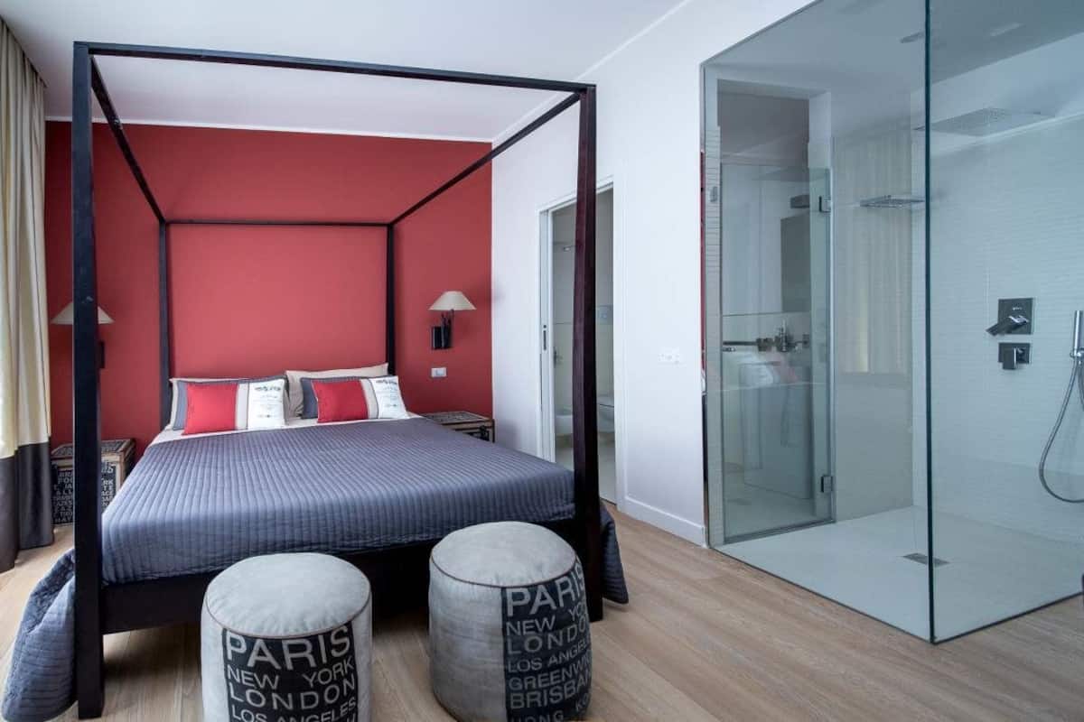 Une chambre moderne avec un lit à baldaquin, une cabine de douche en verre et deux poufs ornés de noms de villes.
