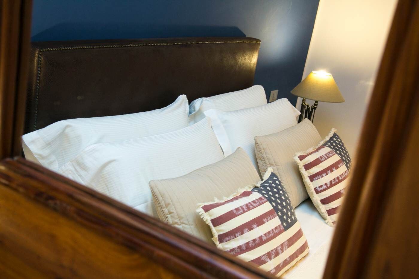 A neatly made bed with white linens and two pillows, one with an American flag design, in a room with a dark headboard and a bedside lamp.