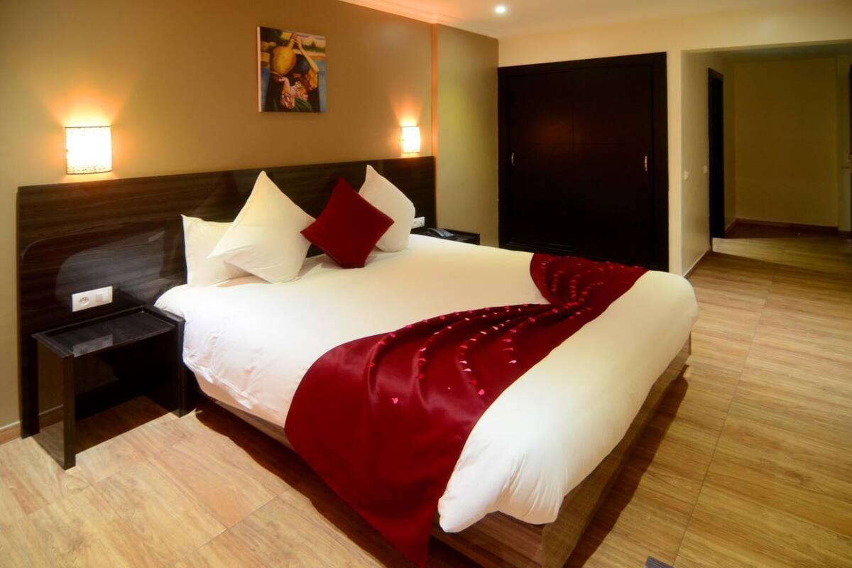 Quarto de hotel com uma cama grande, duas mesas de cabeceira, um criado-mudo, um cobertor vermelho e uma pintura na parede.