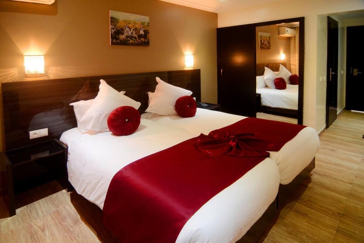 Quarto de hotel com uma cama grande, duas mesas de cabeceira, um criado-mudo, um espelho e um quadro na parede.