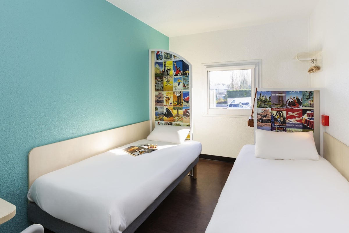 A hotel room with two single beds, a window with a view, and a wall decorated with travel posters.