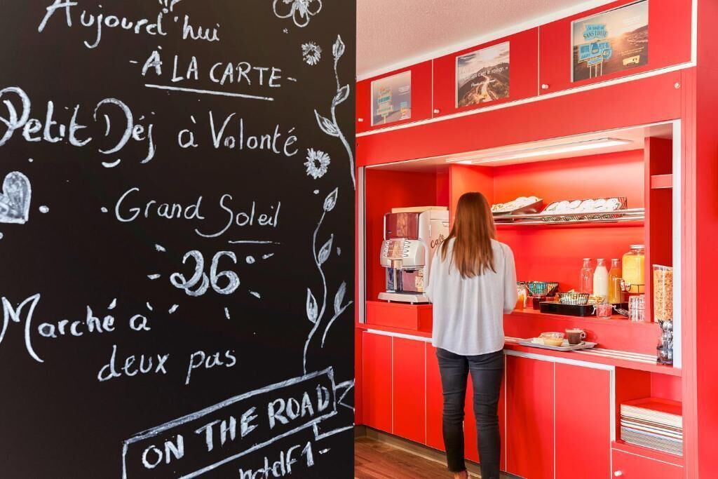 Une personne se tient derrière un comptoir, devant un tableau noir affichant le menu, dans un décor aux couleurs rouge et blanche.