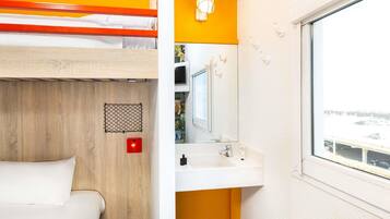 Una cama individual con cabecera de madera, un lavabo blanco con espejo y una ventana con vistas a un aparcamiento.