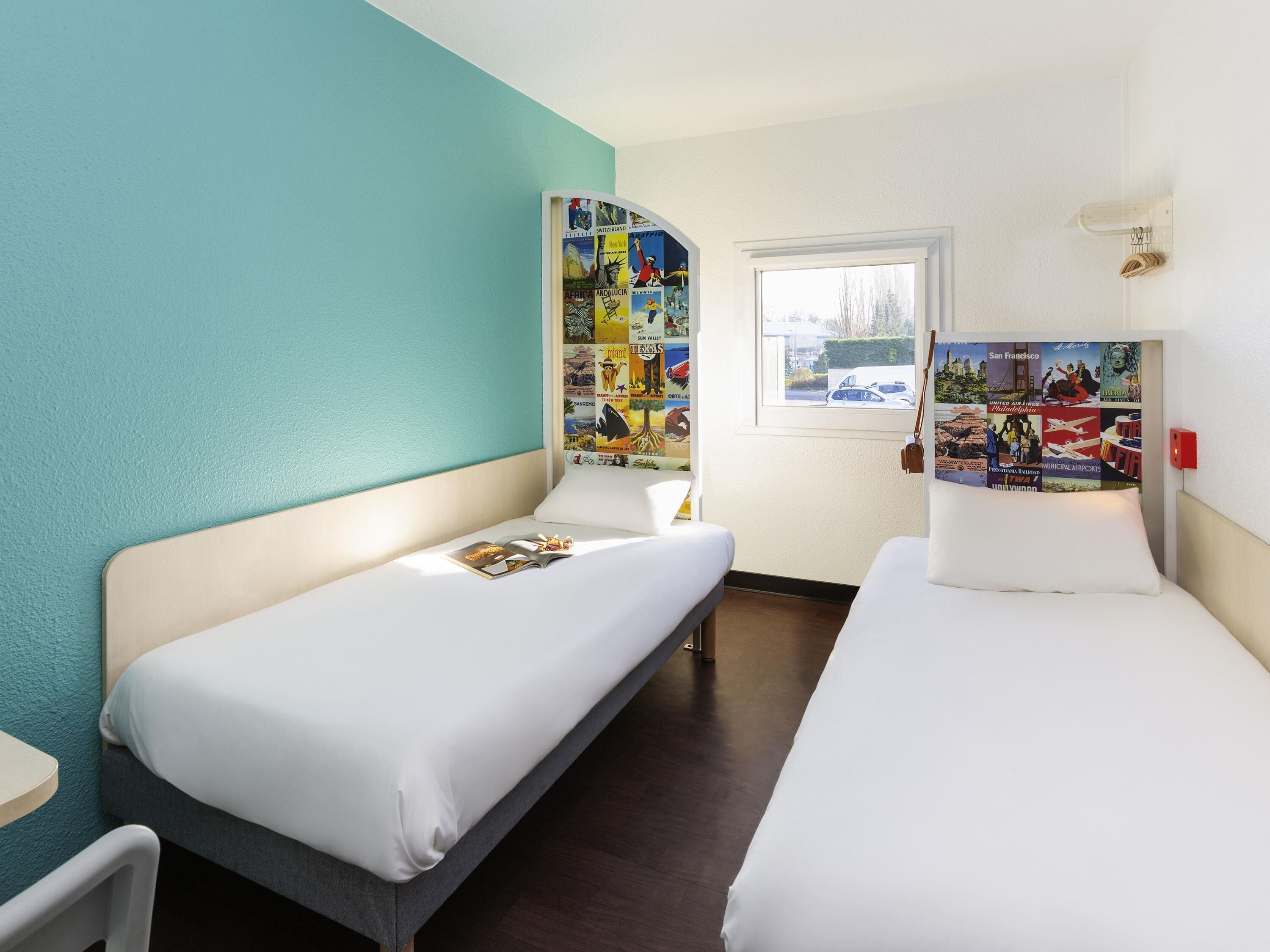 Habitación de hotel con dos camas, una ventana y una pared decorada con carteles de viajes.