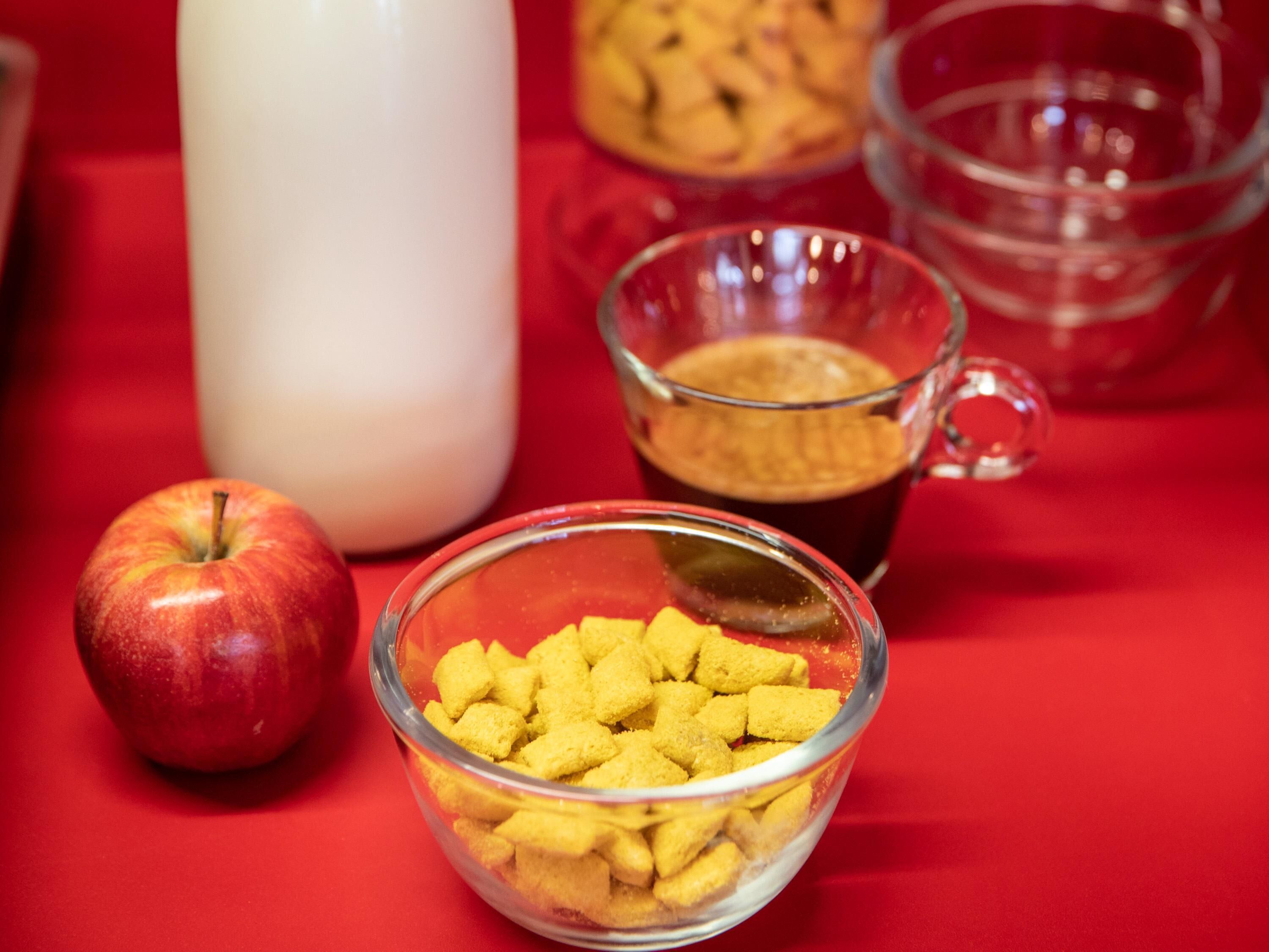 Una mesa roja con un bol de cristal lleno de cubos amarillos, una manzana roja, una taza de cristal y una botella blanca.