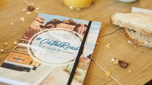 Une table en bois sur laquelle sont disposés des aliments pour le petit-déjeuner, notamment des toasts, des céréales et du jus d’orange. On peut également apercevoir un livre intitulé « Sur la route ».