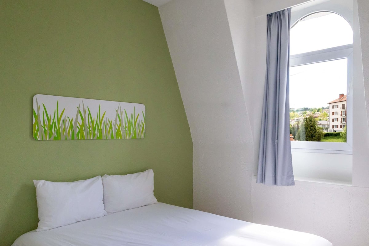 Une chambre à coucher avec un lit, un mur vert, une fenêtre donnant sur des immeubles et un tableau représentant de l’herbe.