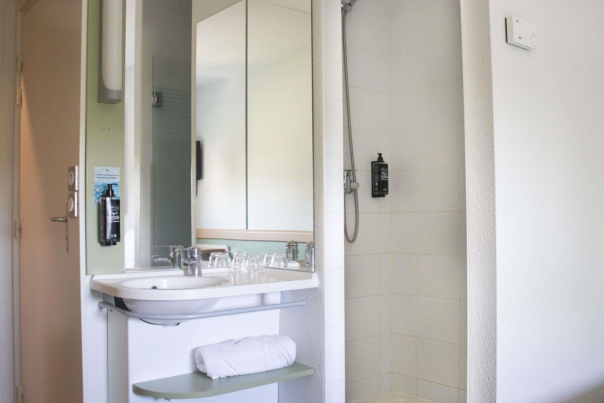 Un baño con lavamanos, espejo, toallero, ducha y dispensador de jabón.
