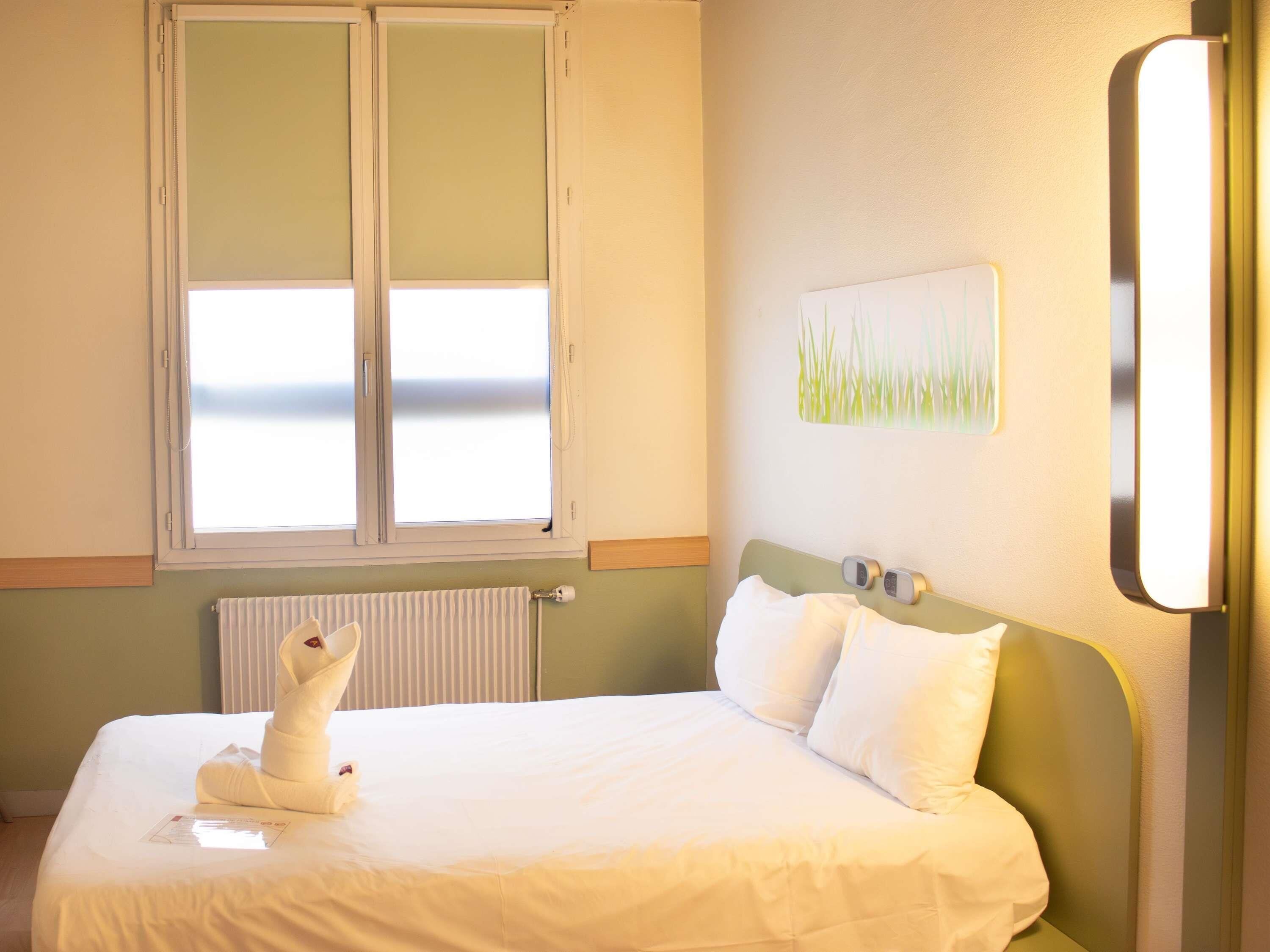 A hotel room with a bed, a window, a radiator, and a wall-mounted light fixture.