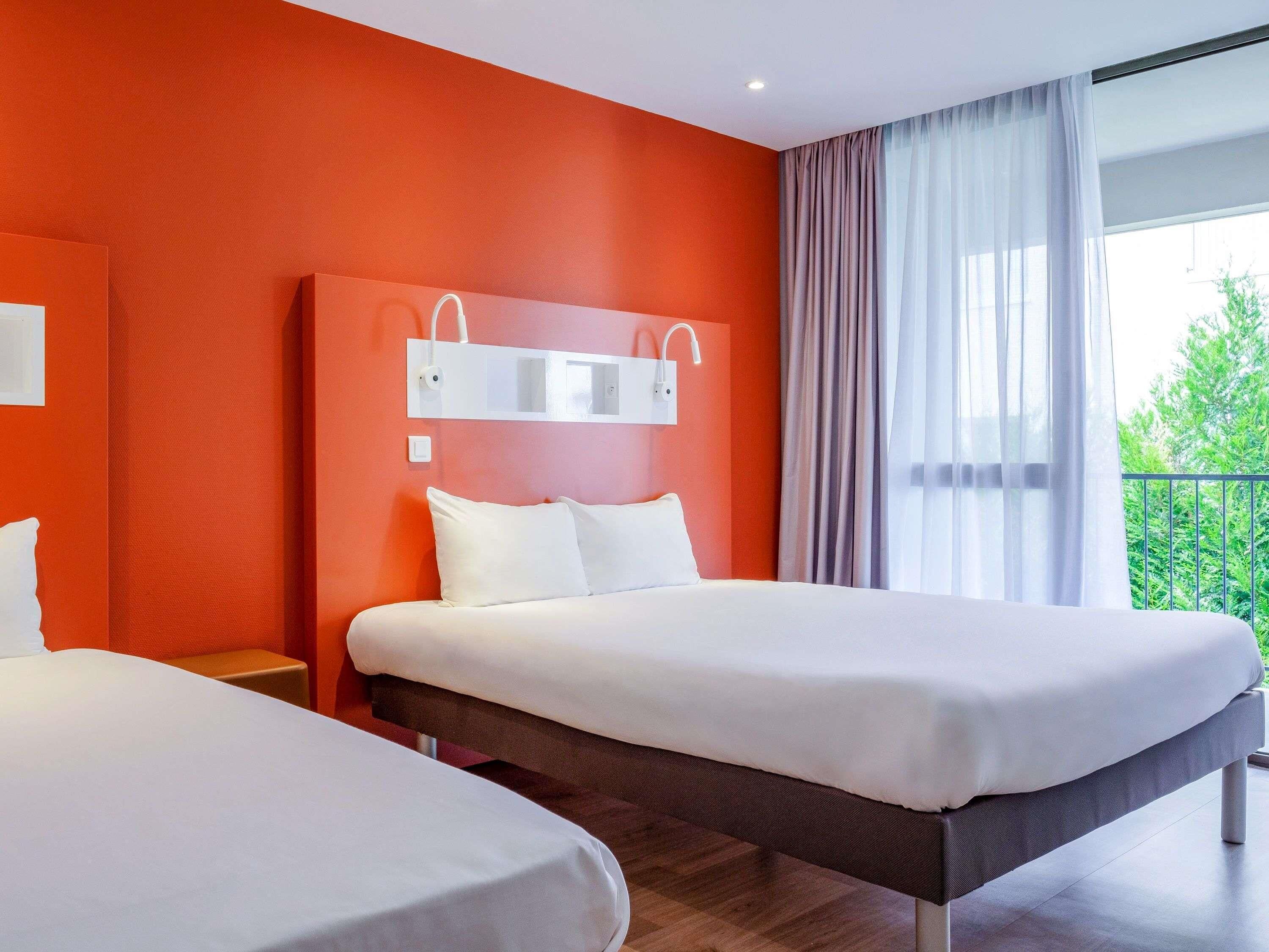 Une chambre d’hôtel avec deux lits, une grande fenêtre avec des rideaux et un balcon agrémenté de plantes.