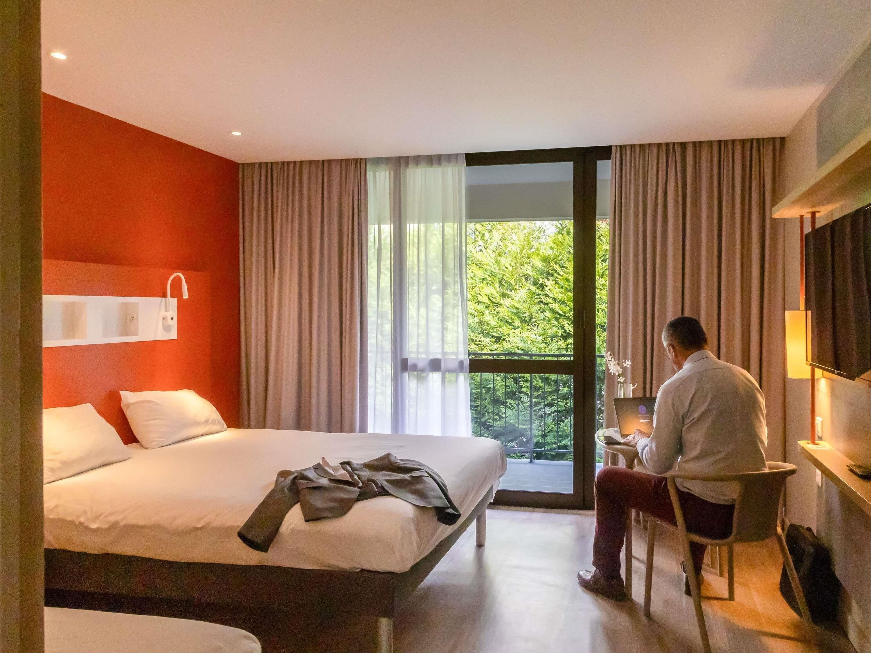 Habitación de hotel con cama, escritorio, silla, televisor y balcón con cortinas.
