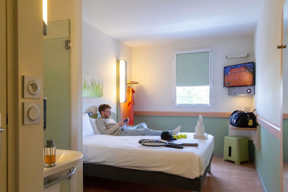 Une chambre d’hôtel avec un lit, un téléviseur fixé au mur, une fenêtre et un petit tabouret.
