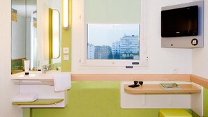 Un baño moderno con un espejo grande, un lavamanos, una ventana con vistas a edificios y un televisor instalado en la pared.