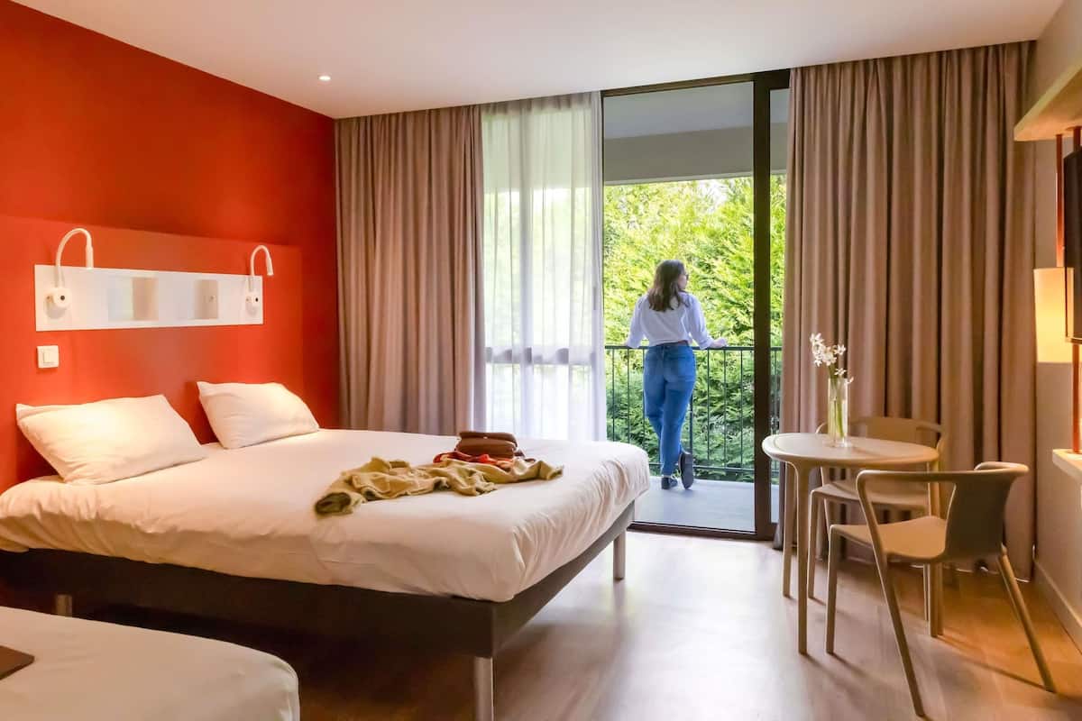 Une chambre d’hôtel avec un lit, une petite table avec des chaises, un balcon donnant sur un espace verdoyant et une fenêtre avec des rideaux.