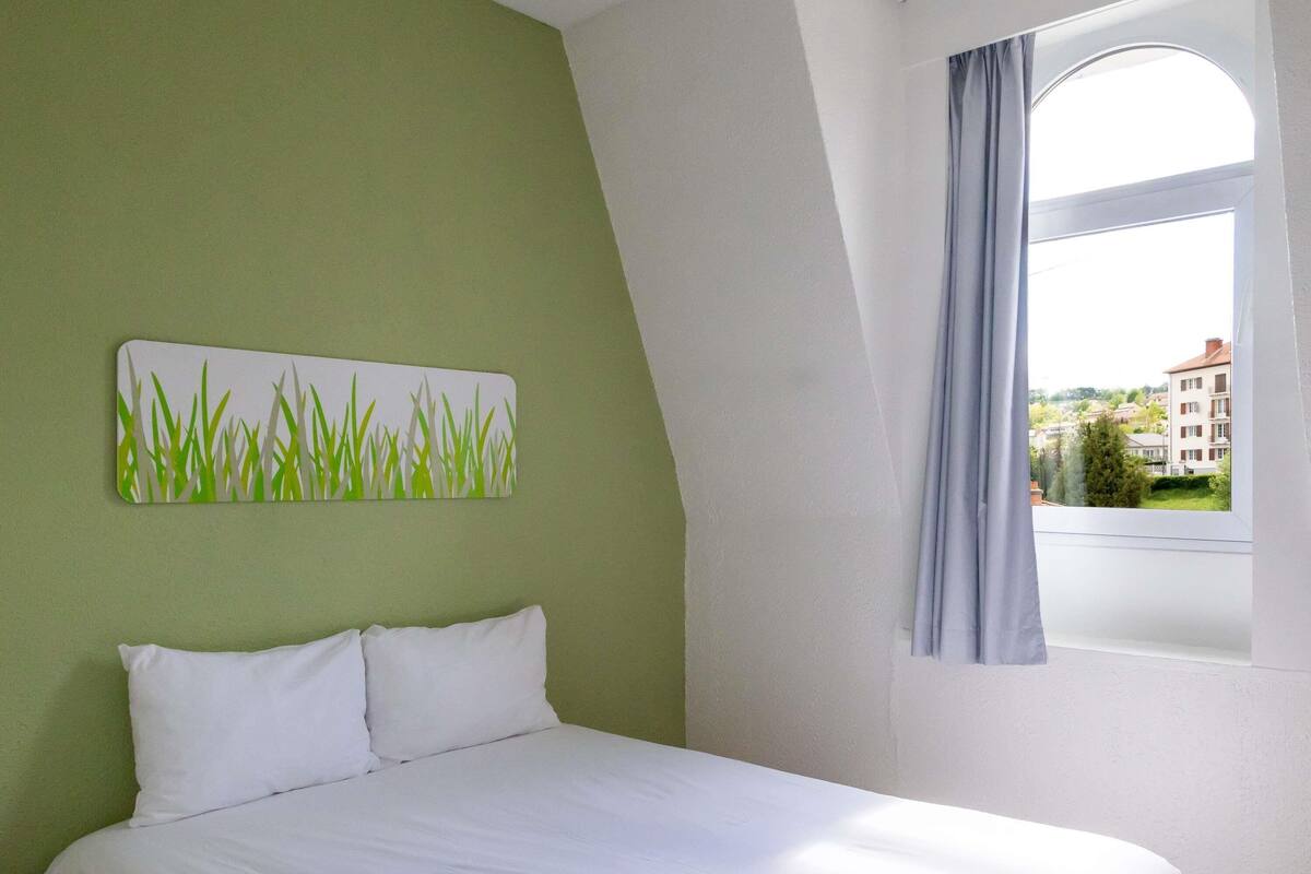 A bedroom with a bed, a green wall, a window with curtains, and a view of buildings outside.