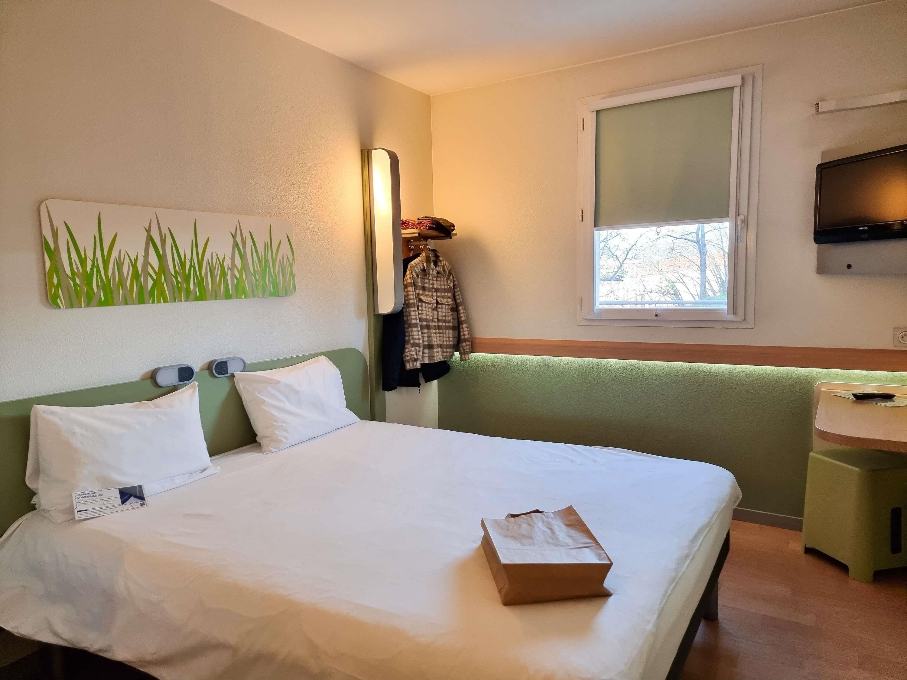 Een hotelkamer met een bed, nachtkastje, televisie, raam en een spiegel aan de muur.