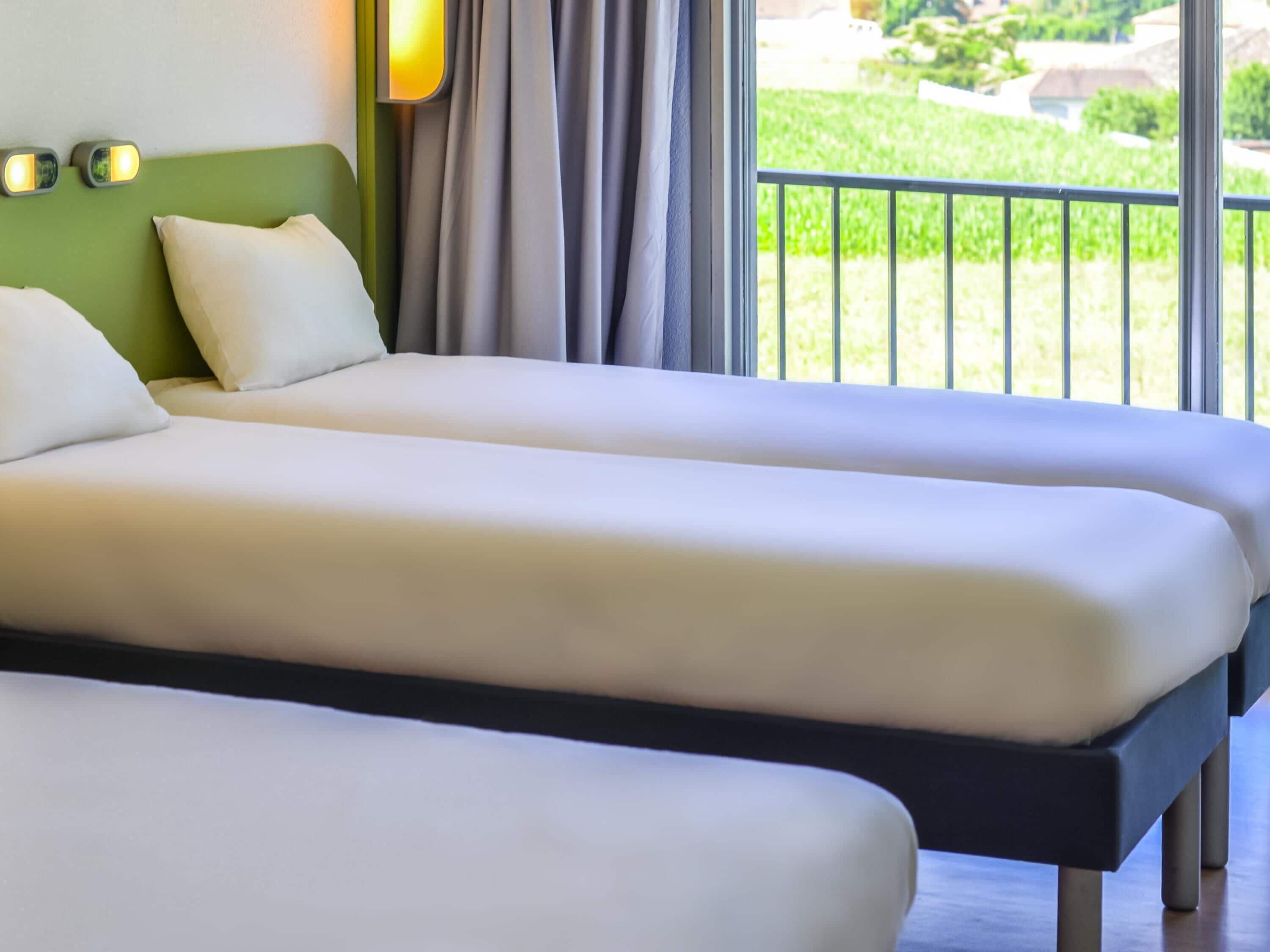 Een hotelkamer met twee bedden, een groen hoofdeinde en uitzicht op een tuin vanaf een balkon.