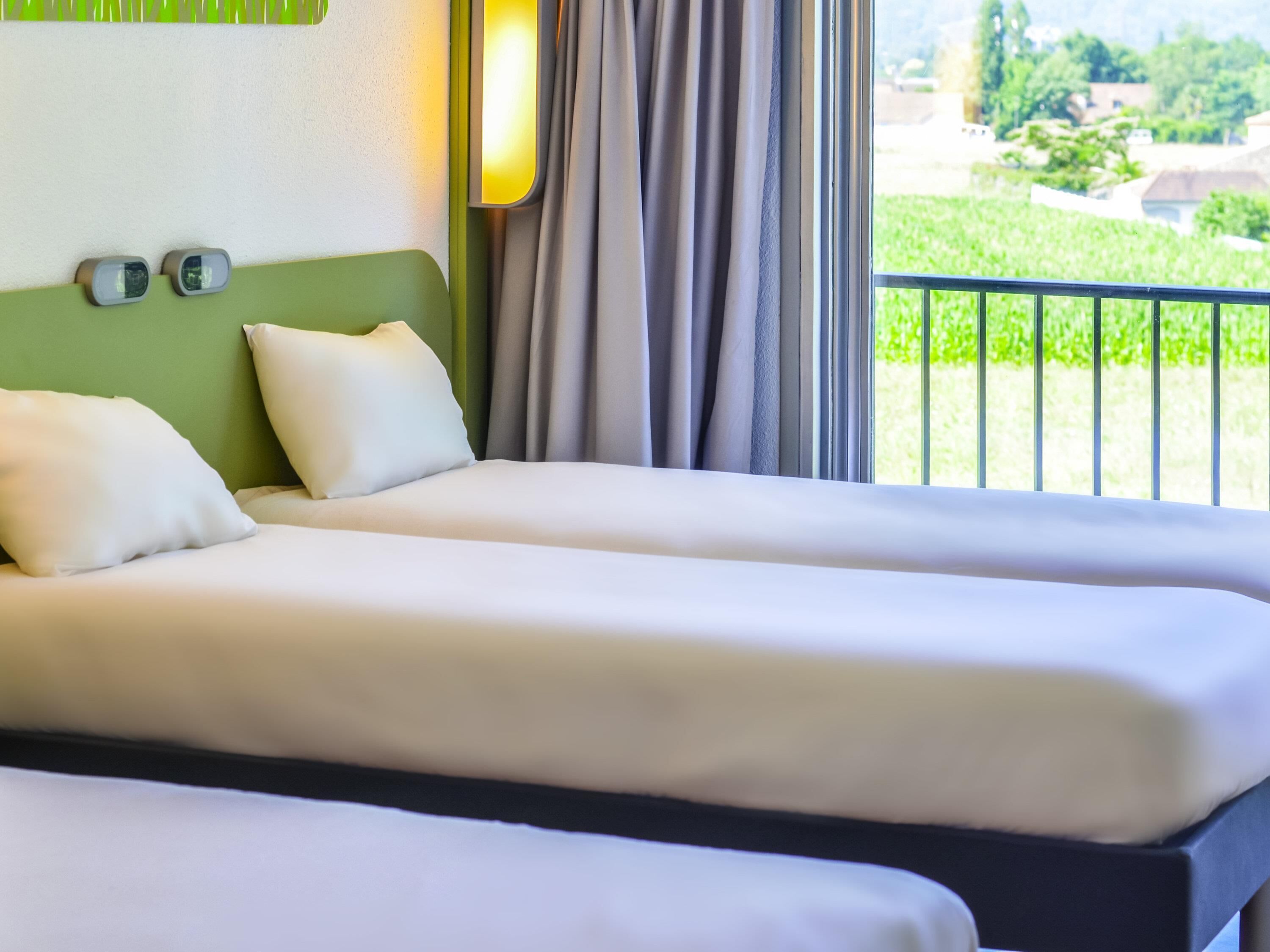 A hotel room with two beds, a green headboard, and a view of a garden through a balcony.