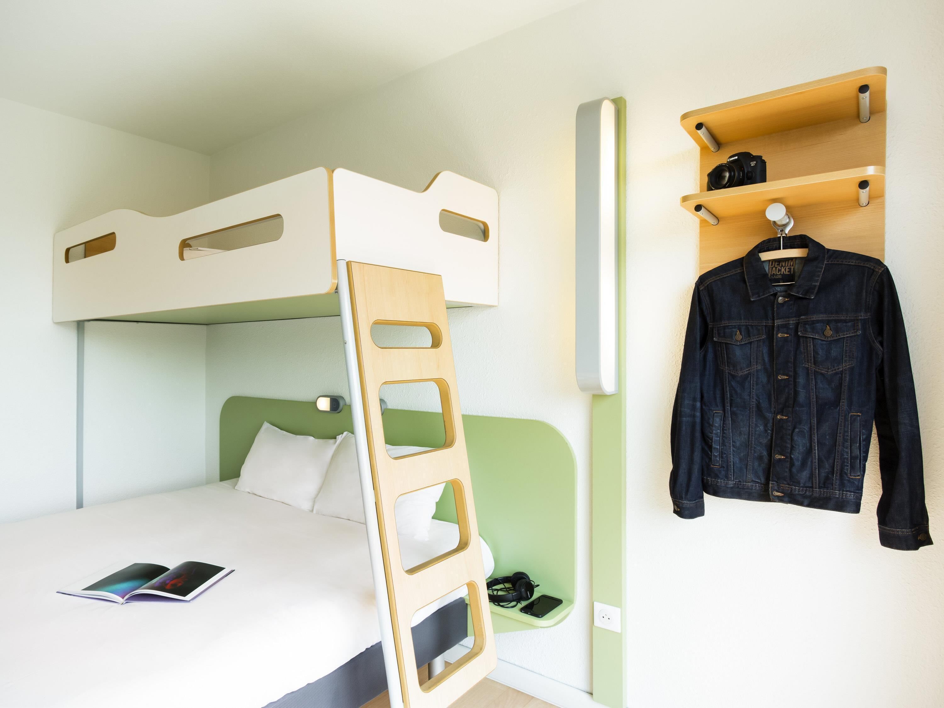 Una cama litera con escalera, una mesita de noche con un libro, auriculares, un teléfono inteligente y un perchero con una chaqueta vaquera.