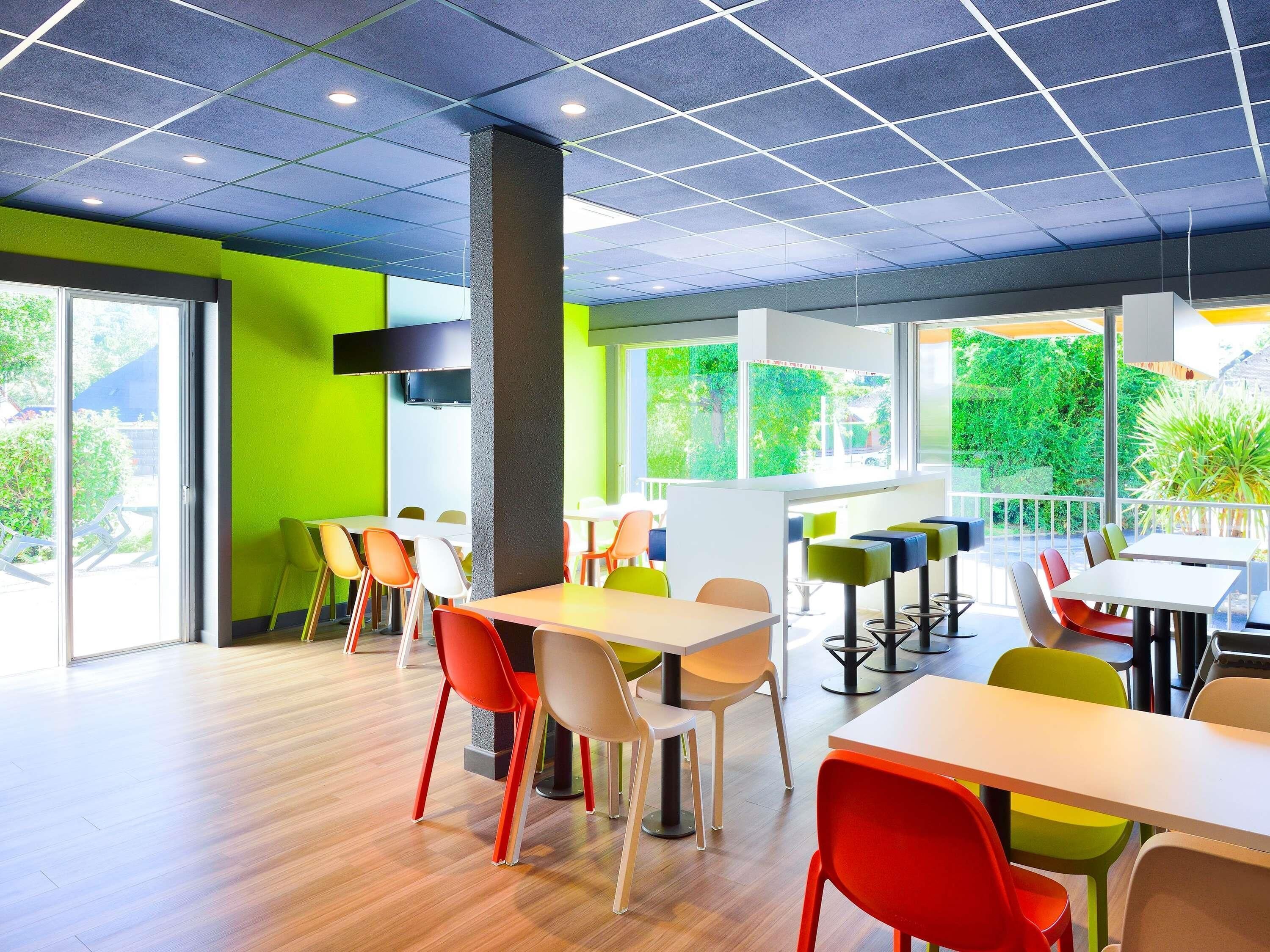 A modern dining area with tables and chairs, large windows, and a view of greenery outside.