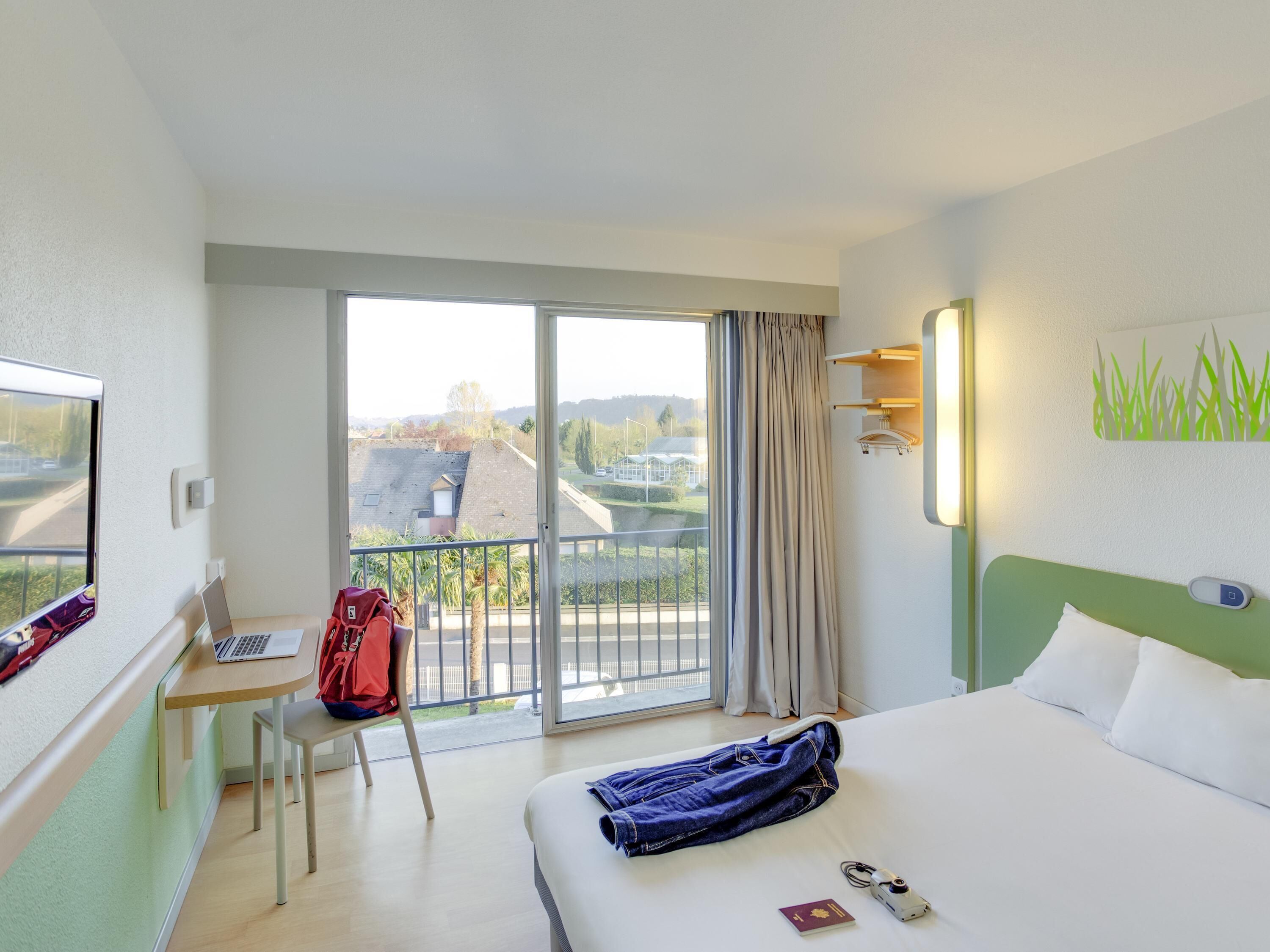 A hotel room with a bed, a desk with a laptop, a chair, a balcony with a view, and a window with curtains.