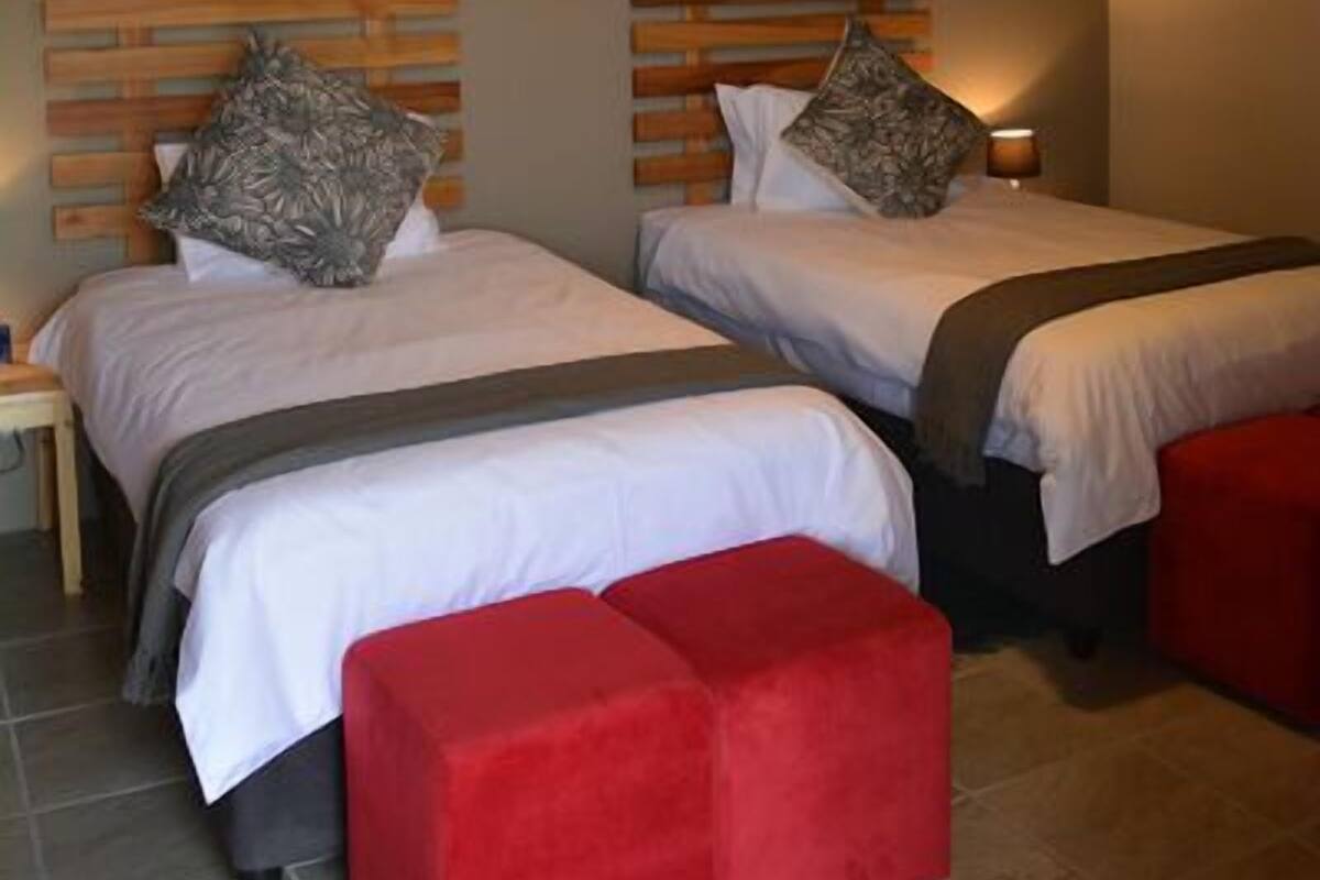 Deux lits simples avec des têtes de lit en bois, un pouf rouge, un luminaire suspendu et un appareil de climatisation fixé au mur.