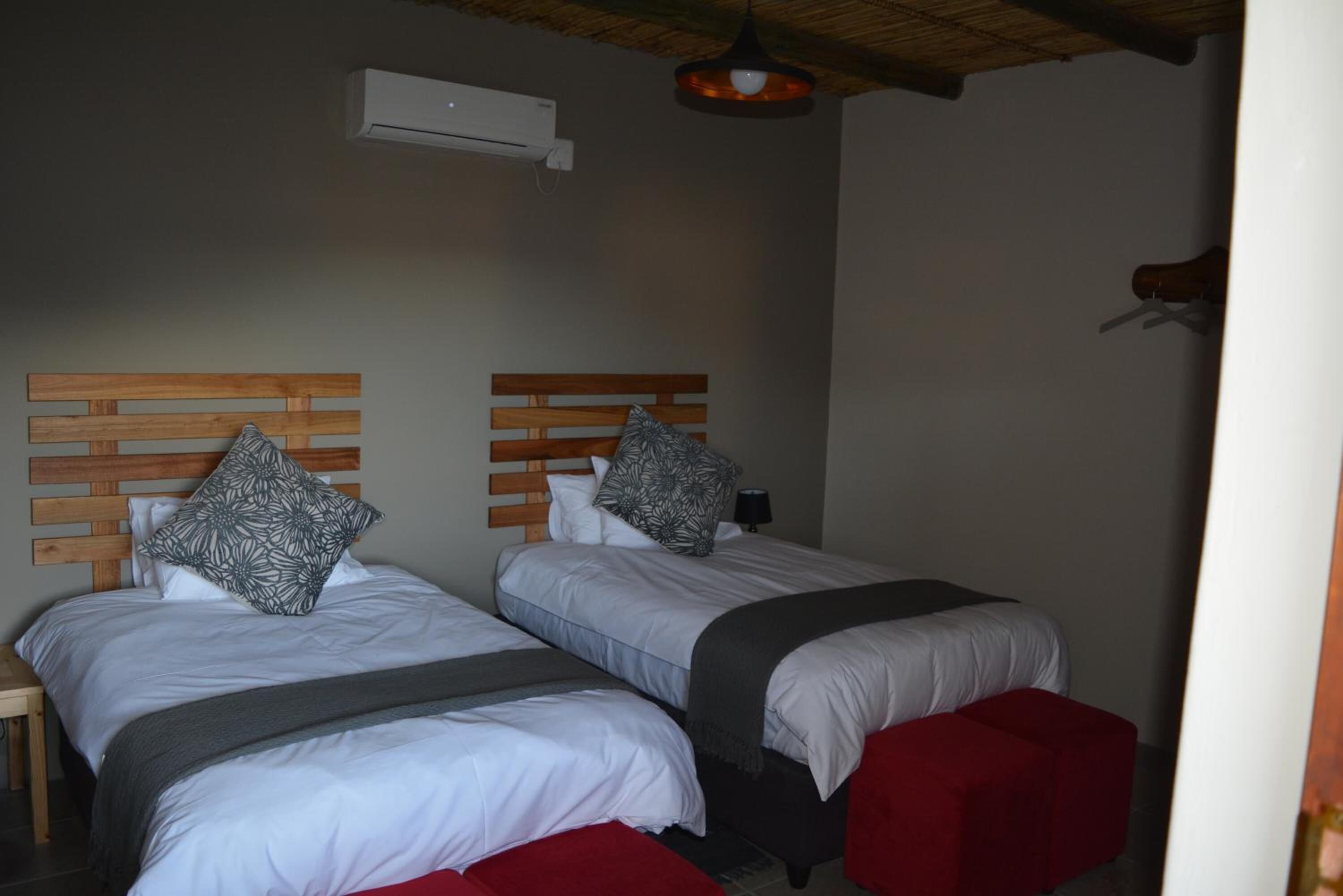 Habitación con dos camas, cada una con cabecero de madera, ropa blanca y cojines rojos. Hay una mesita y un aire acondicionado de pared.
