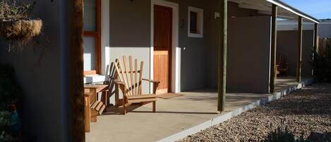 Un porche cubierto con sillas y una mesa de madera, una puerta de madera y una maceta colgante.