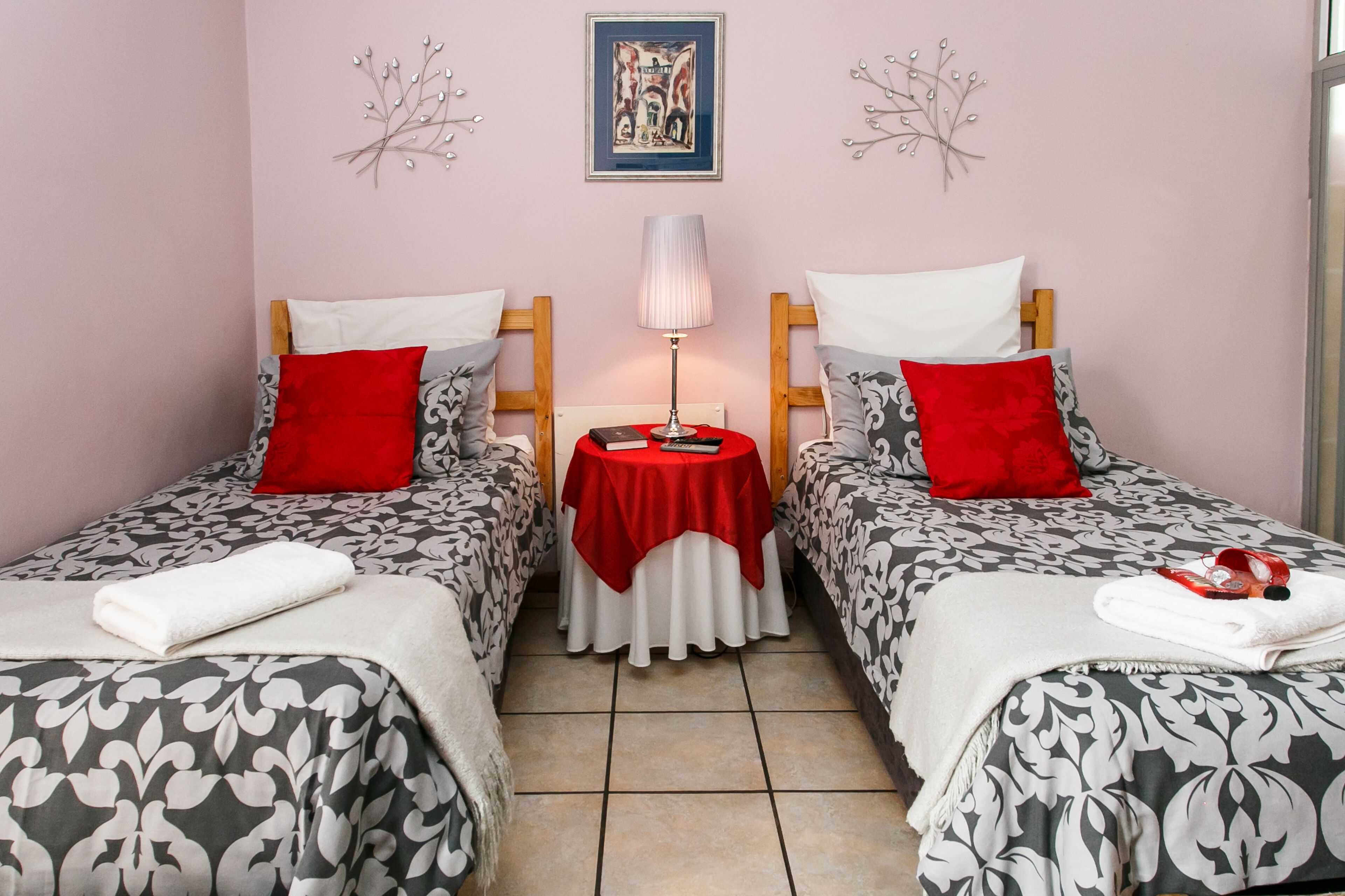 A room with two single beds, each with a red pillow and a white blanket. There is a small table with a red tablecloth in the center, a lamp on it, and a framed picture on the wall.