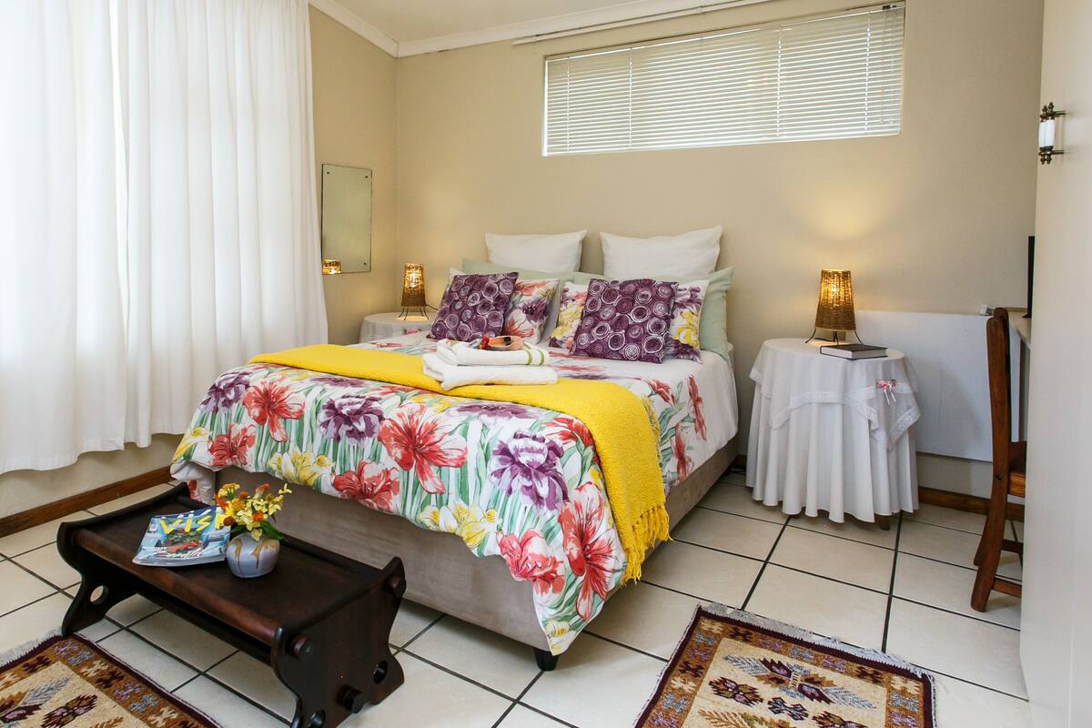 Une chambre à coucher bien rangée, avec une couvre-lit à motifs floraux, une table de chevet avec une lampe et une petite table sur laquelle sont posés un magazine et un bouquet de fleurs.