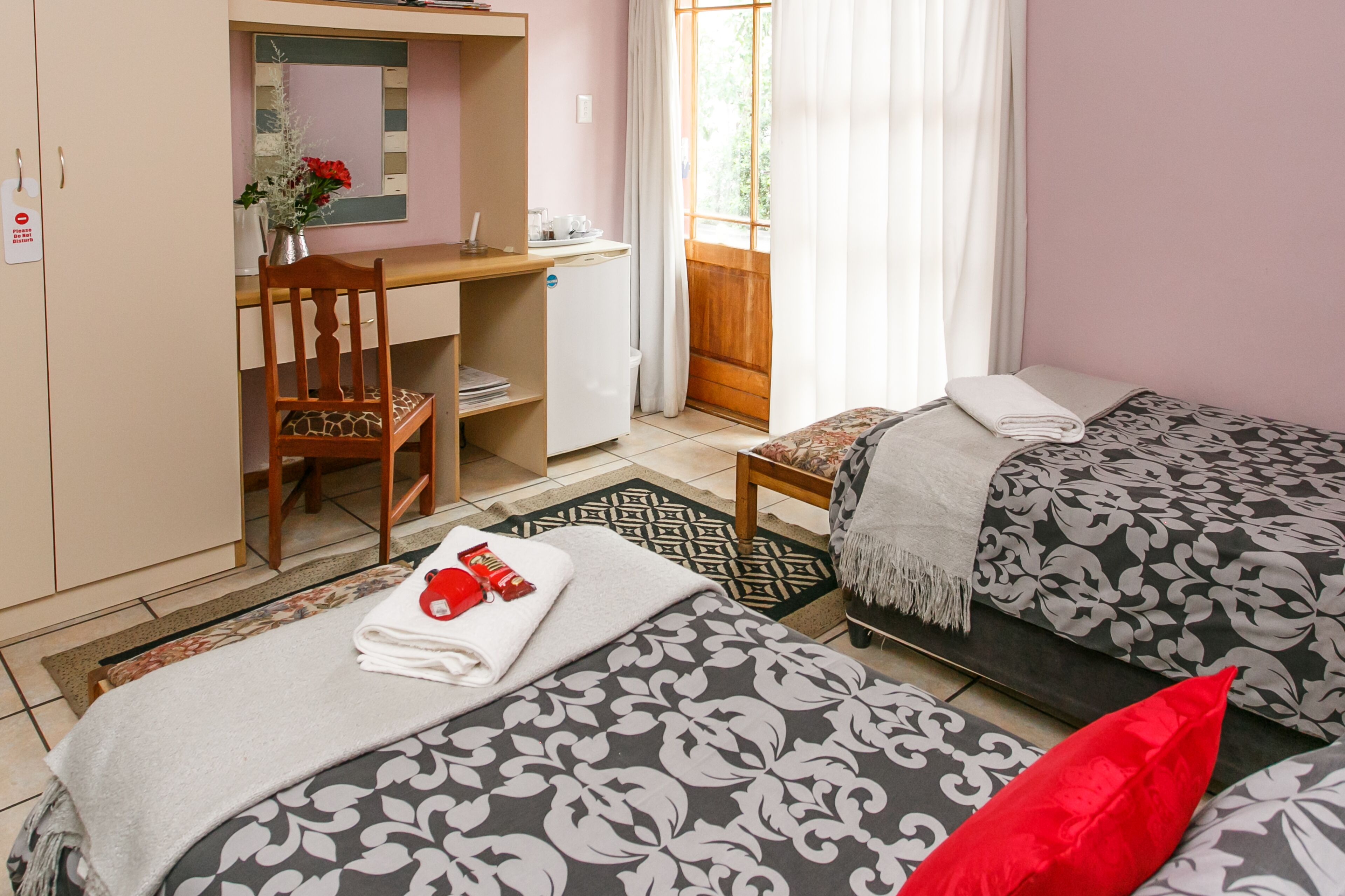 Una habitación con dos camas, un escritorio, una silla, un refrigerador pequeño y una ventana con cortinas.