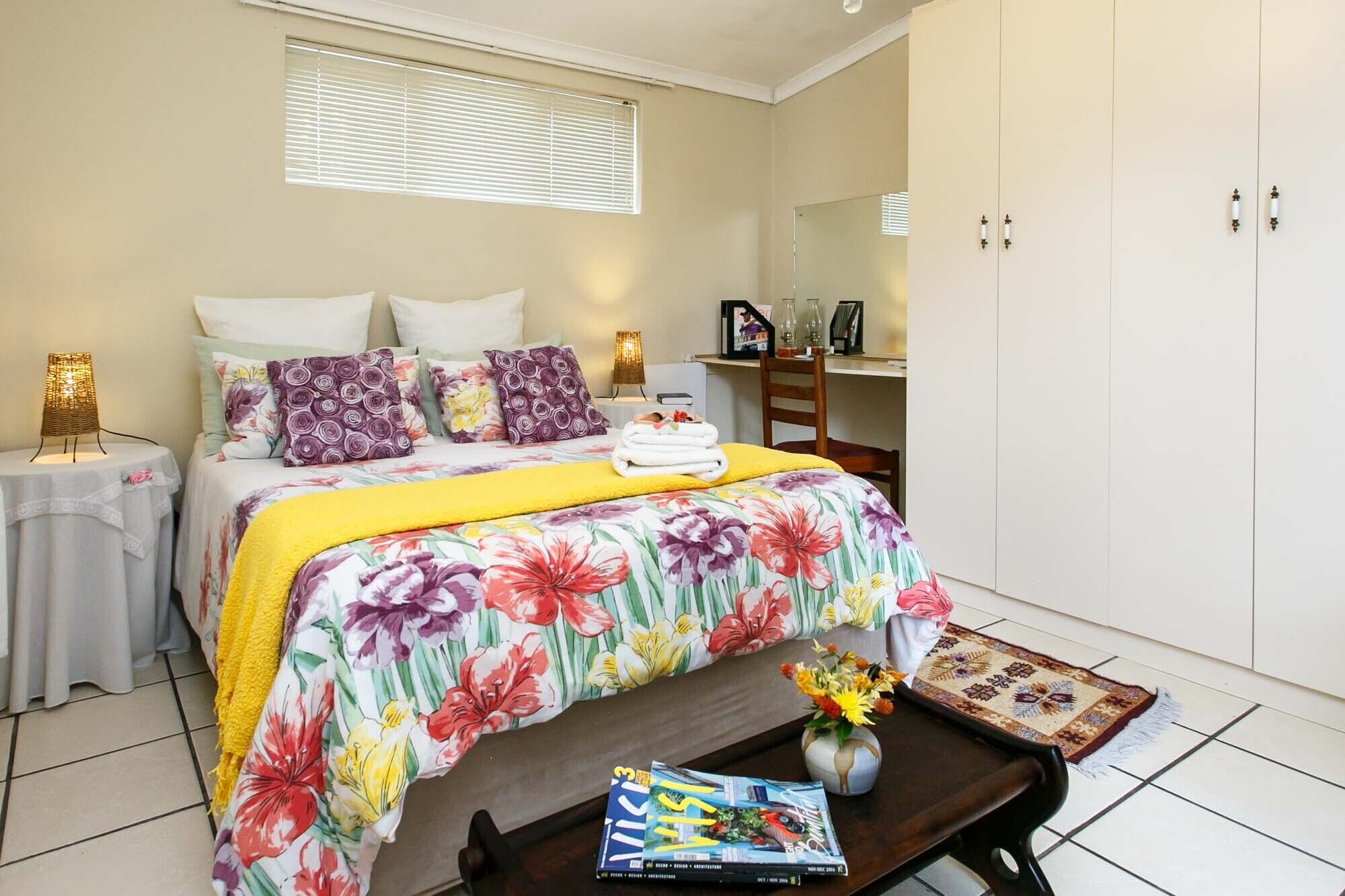 Quarto com cama, mesas de cabeceira, escrivaninha, guarda-roupa e janela com persianas.